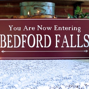 May include: A maroon wooden sign with white text that reads "You Are Now Entering BEDFORD FALLS". The sign has a decorative flourish on each side.