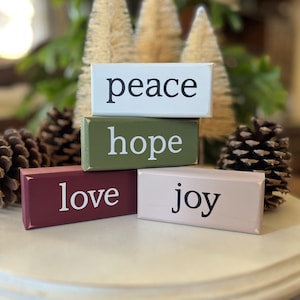 Set of four wood Christmas stacking blocks with words peace, hope, love and joy. Sitting on a table riser with bottle trees and pinecones.  blocks are white, tan. burgundy and moss green.