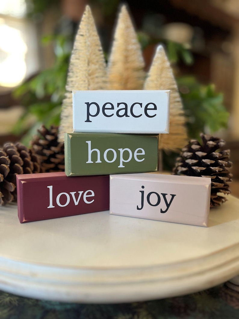 Set of four wood Christmas stacking blocks with words peace, hope, love and joy. Sitting on a table riser with bottle trees and pinecones.  blocks are white, tan. burgundy and moss green.