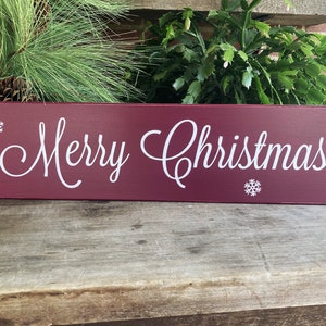May include: A rectangular burgundy wooden sign featuring the white text Merry Christmas in a cursive script font. The sign is decorated with four small white snowflake icons positioned in the corners and near the bottom center.
