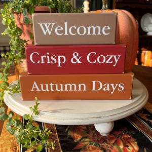 May include: A stack of three rectangular wooden blocks painted in brown, red, and orange with white text. The top block reads Welcome, the middle block reads Crisp & Cozy, and the bottom block reads Autumn Days. The blocks are arranged horizontally.
