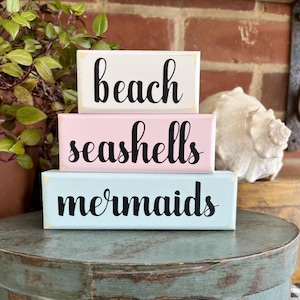May include: Three wooden blocks stacked on top of each other. The top block is white with the word "beach" written in black. The middle block is pink with the word "seashells" written in black. The bottom block is light blue with the word "mermaids" written in black.