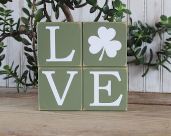 May include: Four green wooden blocks spell the word "LOVE". The letters are white, and one block features a white shamrock. The blocks are arranged to form a square, with a wooden surface in the foreground and greenery in the background.