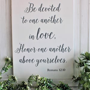 May include: White wooden sign with black calligraphy text that reads "Be devoted to one another in love, Honor one another above yourselves. Romans 12:10".
