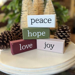 May include: Four rectangular wooden blocks stacked and arranged, featuring the words peace, hope, love, and joy printed in black or white. The blocks are painted in white, olive green, deep red, and light pink with distressed edges.