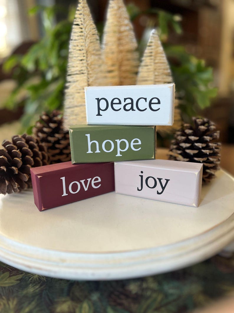 May include: Four rectangular wooden blocks stacked and arranged, featuring the words peace, hope, love, and joy printed in black or white. The blocks are painted in white, olive green, deep red, and light pink with distressed edges.