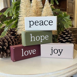 May include: Four rectangular wooden blocks stacked and arranged, featuring the words peace, hope, love, and joy in black or white lettering. The blocks are painted in white, olive green, deep burgundy, and light pink with distressed edges.