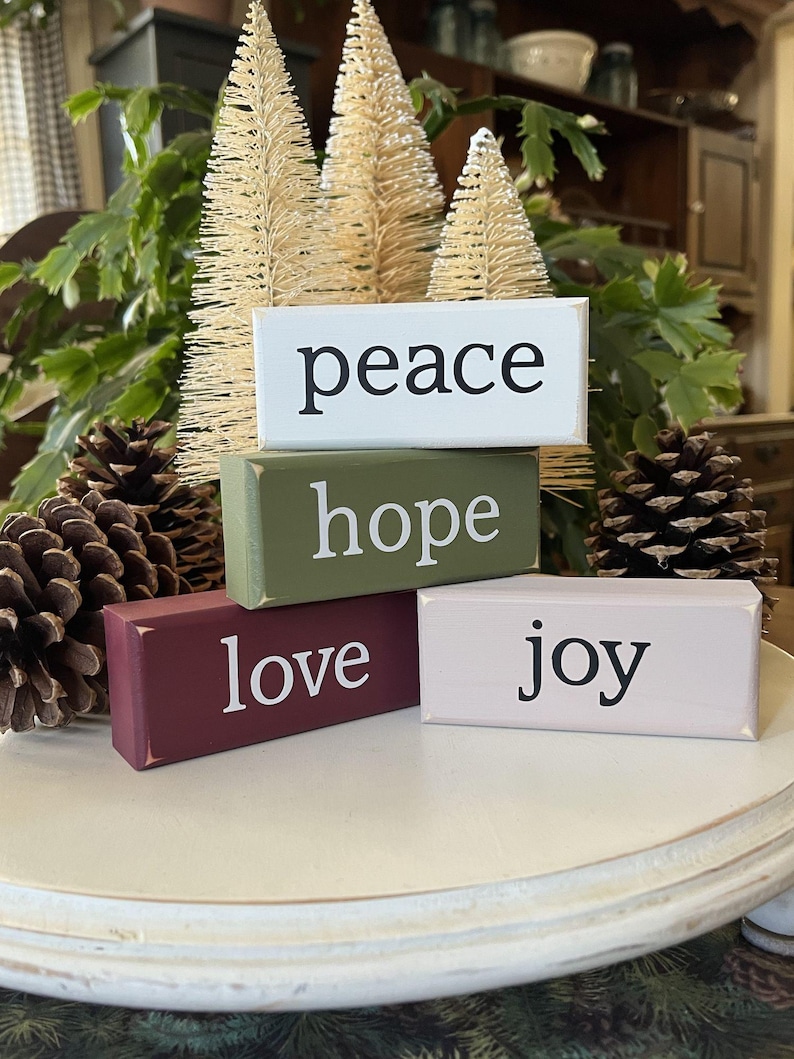 May include: Four rectangular wooden blocks stacked and arranged, featuring the words peace, hope, love, and joy in black or white lettering. The blocks are painted in white, olive green, deep burgundy, and light pink with distressed edges.
