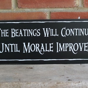 May include: Black wooden sign with white text that reads "The beatings will continue until morale improves."