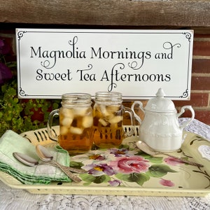 May include: A white tray with a floral design and two mason jars filled with iced tea. A white sugar dispenser and two spoons are also on the tray. A sign above the tray reads "Magnolia Mornings and Sweet Tea Afternoons".