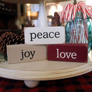 May include: A decorative wooden tray holds three rectangular blocks painted white, tan, and burgundy, each with a single word in black: "peace", "joy", and "love". A blue glass jar filled with candy canes and a pinecone are also on the tray.