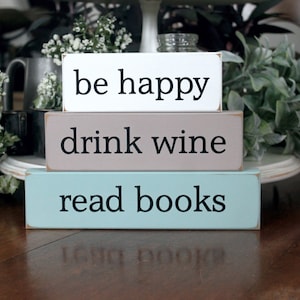 May include: Three wooden signs stacked on top of each other. The top sign is white with black text that reads "be happy". The middle sign is brown with black text that reads "drink wine". The bottom sign is light blue with black text that reads "read books".
