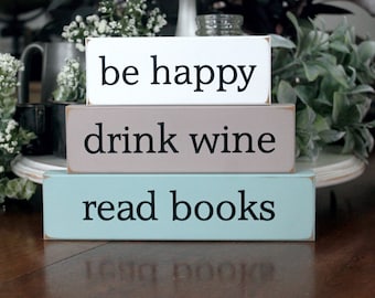 May include: Three wooden signs stacked on top of each other. The top sign is white with black text that reads "be happy". The middle sign is brown with black text that reads "drink wine". The bottom sign is light blue with black text that reads "read books".
