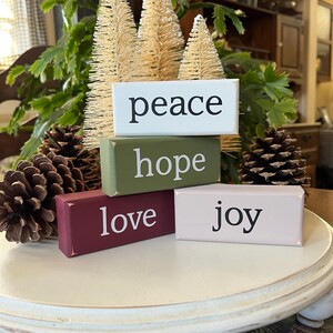 May include: Four rectangular wooden blocks stacked and arranged, featuring the words peace, hope, love, and joy in black or white lettering. The blocks are painted in white, olive green, deep red, and light pink, accompanied by three pinecones.