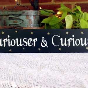 May include: Black wooden sign with white lettering that reads "Curiouser & Curiouser" with gold stars on a black background.