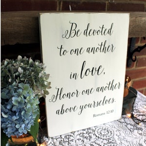 May include: White wooden sign with black calligraphy text that reads "Be devoted to one another in love. Honor one another above yourselves. Romans 12:10".