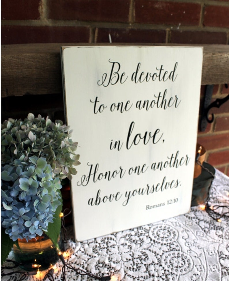 May include: White wooden sign with black calligraphy text that reads "Be devoted to one another in love. Honor one another above yourselves. Romans 12:10".