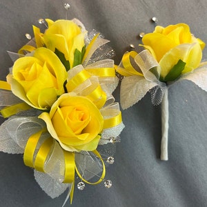 May include: A yellow rose corsage and boutonniere set with white tulle and yellow ribbon. The corsage has three roses and the boutonniere has one rose.