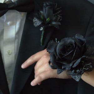 May include: A black rose wrist corsage and boutonniere set. The wrist corsage features a large black rose with decorative accents. The boutonniere is a smaller black rose with green accents. The set is displayed on a black suit.