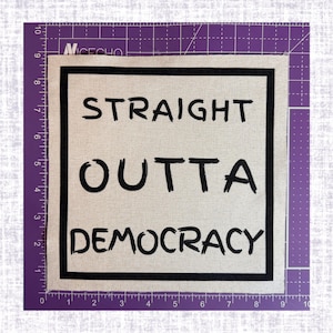 May include: A square fabric patch with a beige background and a black border. The text "STRAIGHT OUTTA DEMOCRACY" is printed in black, hand-drawn style lettering. The patch is placed on a purple cutting mat with markings up to 9 inches.