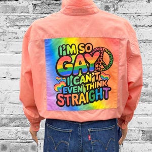 May include: A coral-colored long-sleeve shirt featuring a large square patch on the back with a rainbow tie-dye background. The patch displays the text "I'M SO GAY I CAN'T EVEN THINK STRAIGHT" in black letters, accompanied by a leopard-print peace sign.