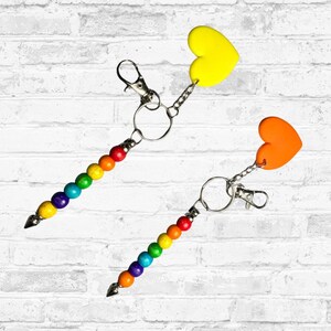 May include: Two keychains with rainbow-colored beads and heart-shaped charms. One keychain has a yellow heart, the other an orange heart. Each keychain has a silver clasp and a silver ring.