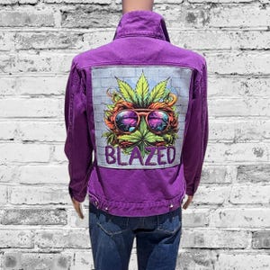 May include: A purple denim jacket with a graphic on the back featuring a cannabis leaf, sunglasses, and the word "BLAZED". The jacket has a button-up front and is worn with blue jeans.