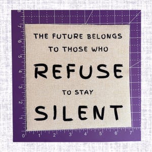 May include: A square beige fabric patch featuring the black embroidered text "THE FUTURE BELONGS TO THOSE WHO REFUSE TO STAY SILENT". The patch is positioned against a purple cutting mat with a visible ruler measuring approximately 7 inches wide.