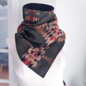 May include: A dark grey cowl scarf with a geometric pattern in shades of red, orange, tan, and navy blue. The scarf is displayed on a mannequin and has a button closure. The inner lining is black.