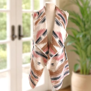 May include: A sleeveless vest featuring an open front design with a draped collar. The garment displays a geometric pattern in shades of cream, dusty rose, brown, and charcoal gray, finished with a visible whipstitch detail along the edges.
