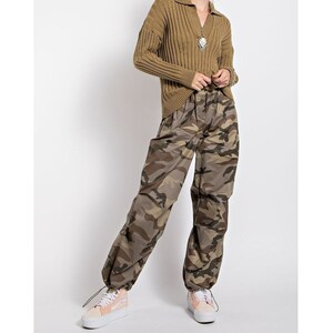 May include: A woman wearing a brown ribbed sweater with a zipper and a pair of camouflage pants. She is wearing pink sneakers.
