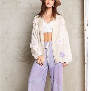May include: A woman wearing a white cardigan with floral embroidery, a white bra, and lavender patchwork pants. She is wearing a wide-brimmed hat and has her hair down.