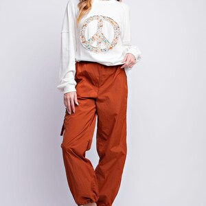 K&ouml;nnte beinhalten: Eine Frau tr&auml;gt ein wei&szlig;es Langarm-Sweatshirt mit einem floralen Peace-Zeichen und rostfarbene Cargohosen. Die Hose hat eine lockere Passform und wird mit wei&szlig;en Turnschuhen kombiniert. Das Outfit ist l&auml;ssig und stilvoll.