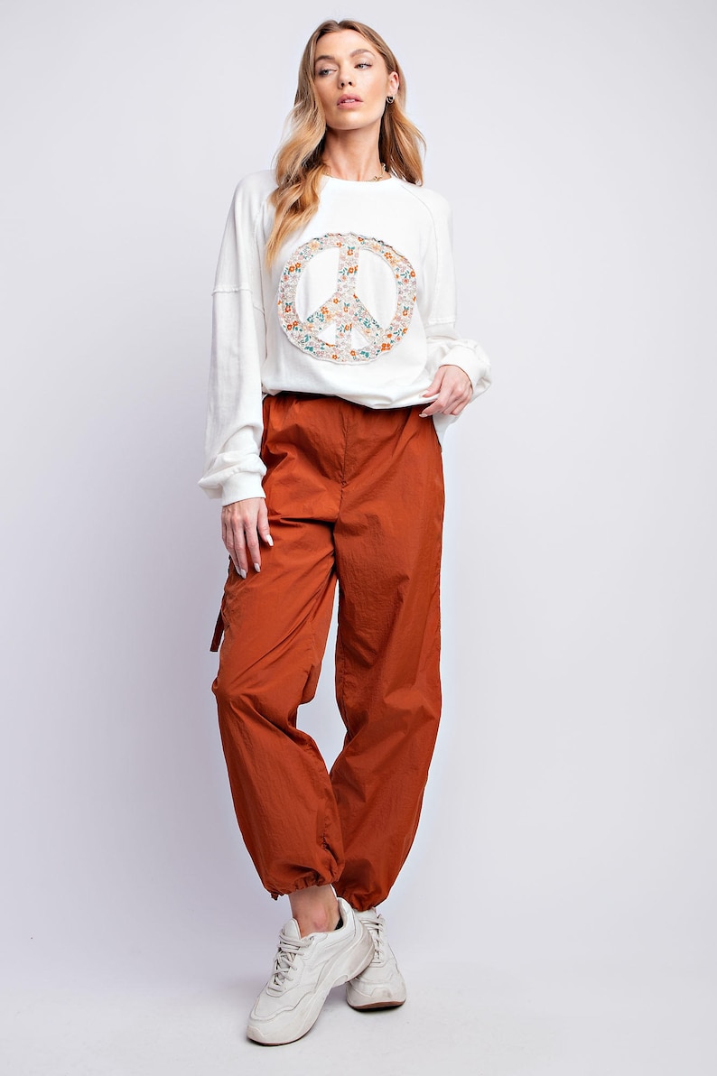 K&ouml;nnte beinhalten: Eine Frau tr&auml;gt ein wei&szlig;es Langarm-Sweatshirt mit einem floralen Peace-Zeichen und rostfarbene Cargohosen. Die Hose hat eine lockere Passform und wird mit wei&szlig;en Turnschuhen kombiniert. Das Outfit ist l&auml;ssig und stilvoll.
