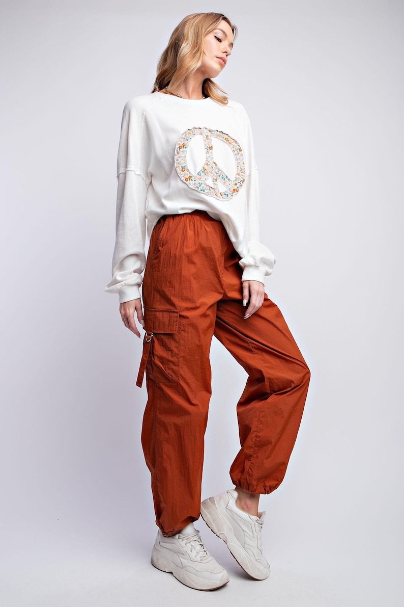 K&ouml;nnte beinhalten: Eine Frau tr&auml;gt ein wei&szlig;es Langarm-Sweatshirt mit einem Peace-Zeichen und rostfarbene Cargohosen. Das Peace-Zeichen ist mit einem floralen Muster gef&uuml;llt. Sie tr&auml;gt auch wei&szlig;e Turnschuhe.