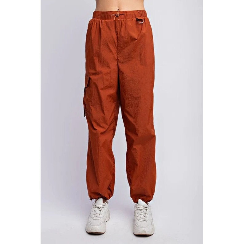 K&ouml;nnte beinhalten: Rostfarbene Cargohose mit elastischem Bund und Kordelzug. Die Hose hat eine lockere Passform, Seitentaschen und elastische B&uuml;ndchen an den Kn&ouml;cheln. Das Model tr&auml;gt wei&szlig;e Turnschuhe. Die Hose ist aus leichtem Material.