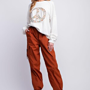 K&ouml;nnte beinhalten: Eine Frau tr&auml;gt ein cremefarbenes Sweatshirt mit einem Peace-Zeichen-Aufdruck und rostfarbene Cargohosen. Die Hose hat Taschen und ist an den Kn&ouml;cheln gerafft. Wei&szlig;e Turnschuhe vervollst&auml;ndigen das Outfit. L&auml;ssiger Stil.