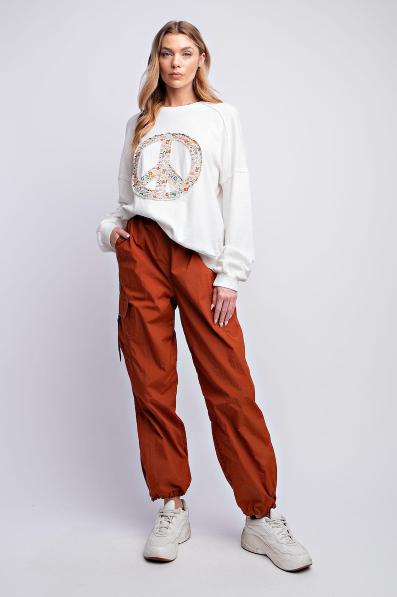 K&ouml;nnte beinhalten: Eine Frau tr&auml;gt ein cremefarbenes Sweatshirt mit einem Peace-Zeichen-Aufdruck und rostfarbene Cargohosen. Die Hose hat Taschen und ist an den Kn&ouml;cheln gerafft. Wei&szlig;e Turnschuhe vervollst&auml;ndigen das Outfit. L&auml;ssiger Stil.