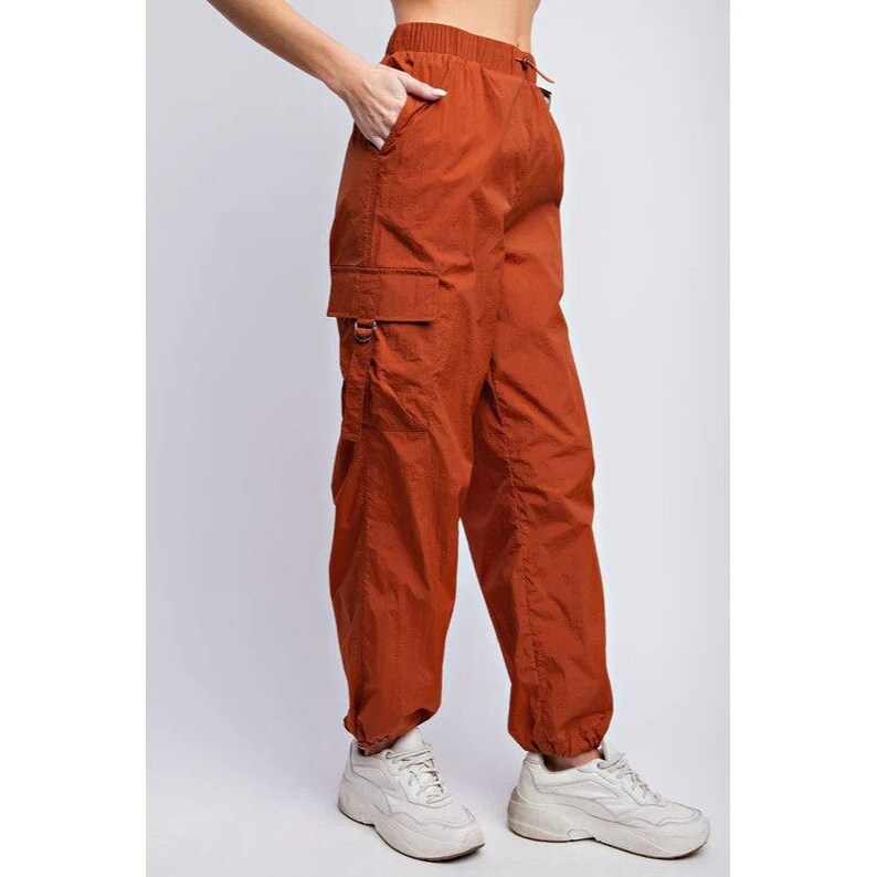 K&ouml;nnte beinhalten: Rostfarbene Cargohose mit Kordelzugbund und elastischen B&uuml;ndchen. Die Hose hat zwei Cargotaschen mit Metallbeschl&auml;gen. Das Model tr&auml;gt wei&szlig;e Turnschuhe. Die Hose ist aus leichtem Material.