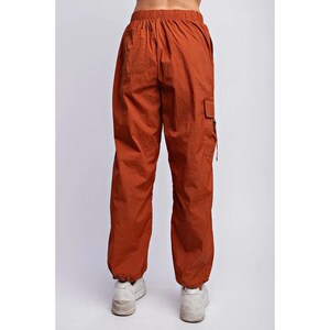 K&ouml;nnte beinhalten: Rostfarbene Cargohose mit elastischem Bund und Kn&ouml;chelb&uuml;ndchen. Die Hose hat eine lockere Passform und eine Cargotasche am rechten Bein. Das Model tr&auml;gt wei&szlig;e Turnschuhe.