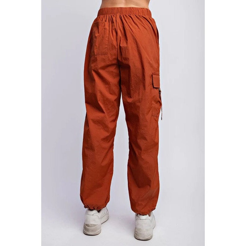 K&ouml;nnte beinhalten: Rostfarbene Cargohose mit elastischem Bund und Kn&ouml;chelb&uuml;ndchen. Die Hose hat eine lockere Passform und eine Cargotasche am rechten Bein. Das Model tr&auml;gt wei&szlig;e Turnschuhe.
