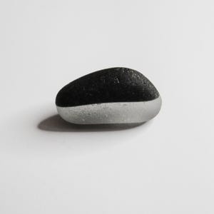 May include: A rounded, oval stone with a two-tone design. The top half is black, the bottom half is light grey. The stone is on a white surface.