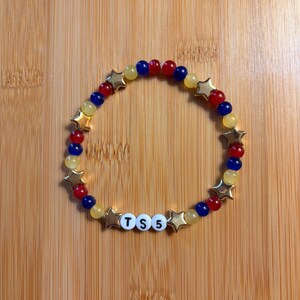 Op de afbeelding: Een cirkelvormige kralenarmband met afwisselend rode, blauwe en gele ronde kralen, afgewisseld met goudkleurige stervormige kralen. De armband bevat drie witte bolvormige kralen met zwarte letters die de tekst TS 5 vormen.