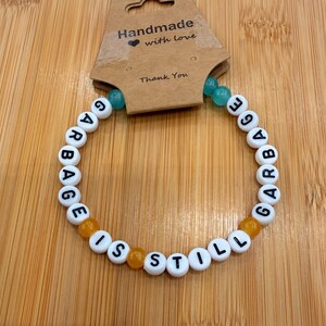 May include: A handmade bracelet with white letter beads spelling "GARBAGE IS STILL GARBAGE". The bracelet features orange and turquoise accent beads. A brown tag above the bracelet reads "Handmade with love" and "Thank You".