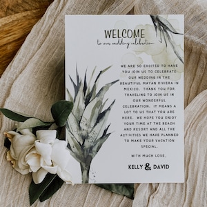 May include: A wedding welcome card with the text "WELCOME to our wedding celebration". The card has watercolor botanical illustrations in green and gray. A white flower and greenery are in the lower left corner.