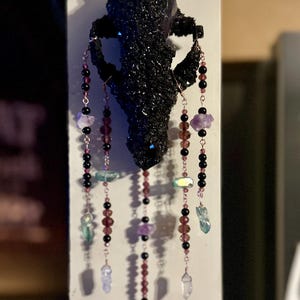 May include: A black, textured skull-shaped wall hanging with dangling strands of beads and crystals. The strands feature black, purple, and clear beads, along with amethyst and quartz crystals. The skull is mounted on a white surface.