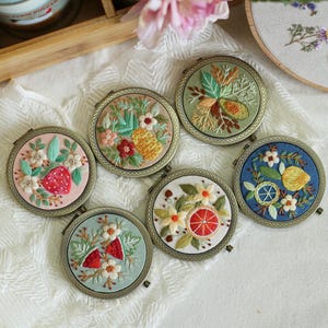 May include: Six round embroidery hoops with different floral designs. Each hoop features a different fruit or flower, including strawberries, pineapples, lemons, and oranges. The hoops are all in a similar style, with a gold metal frame and a white background.
