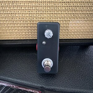 May include: A black guitar effects pedal with a silver footswitch, a white knob, and a red accent. The pedal has a small LED light and is rectangular in shape. The background is a woven tan material.