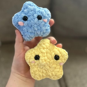 May include: Two handmade, plush star-shaped toys. One is light blue, the other is yellow. Both have black button eyes and pink cheeks. The toys are made of a soft, textured material, and are held in a person's hand.