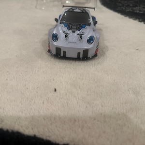 May include: A small, white and blue toy race car with the word "GALLOP" printed on the front. The car has black accents, a rear spoiler, and blue headlights. The toy car is on a white surface.